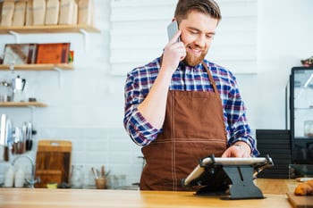 Handsome smiling barista with beard taking order on cell phone and using tablet in cafeteria.jpeg